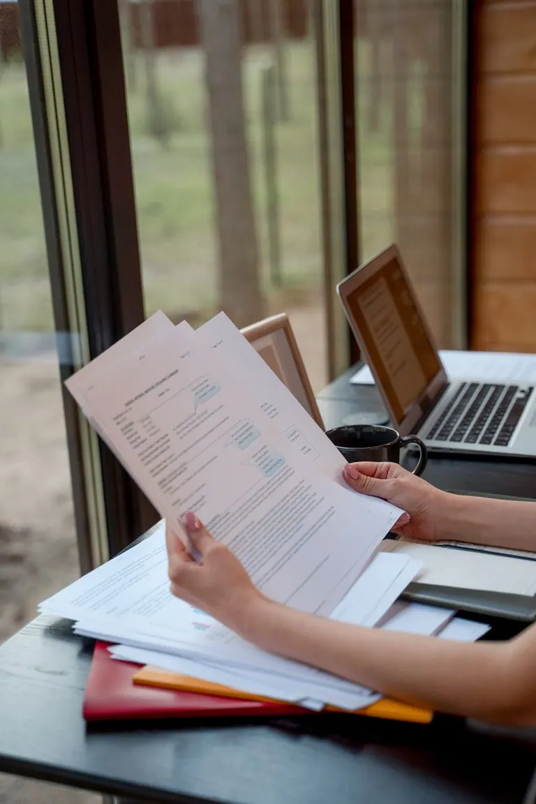 Personne tenant et consultant des documents près d'une fenêtre avec des ordinateurs portables sur un bureau. Personne tenant et consultant des documents près d'une fenêtre avec des ordinateurs portables sur un bureau.